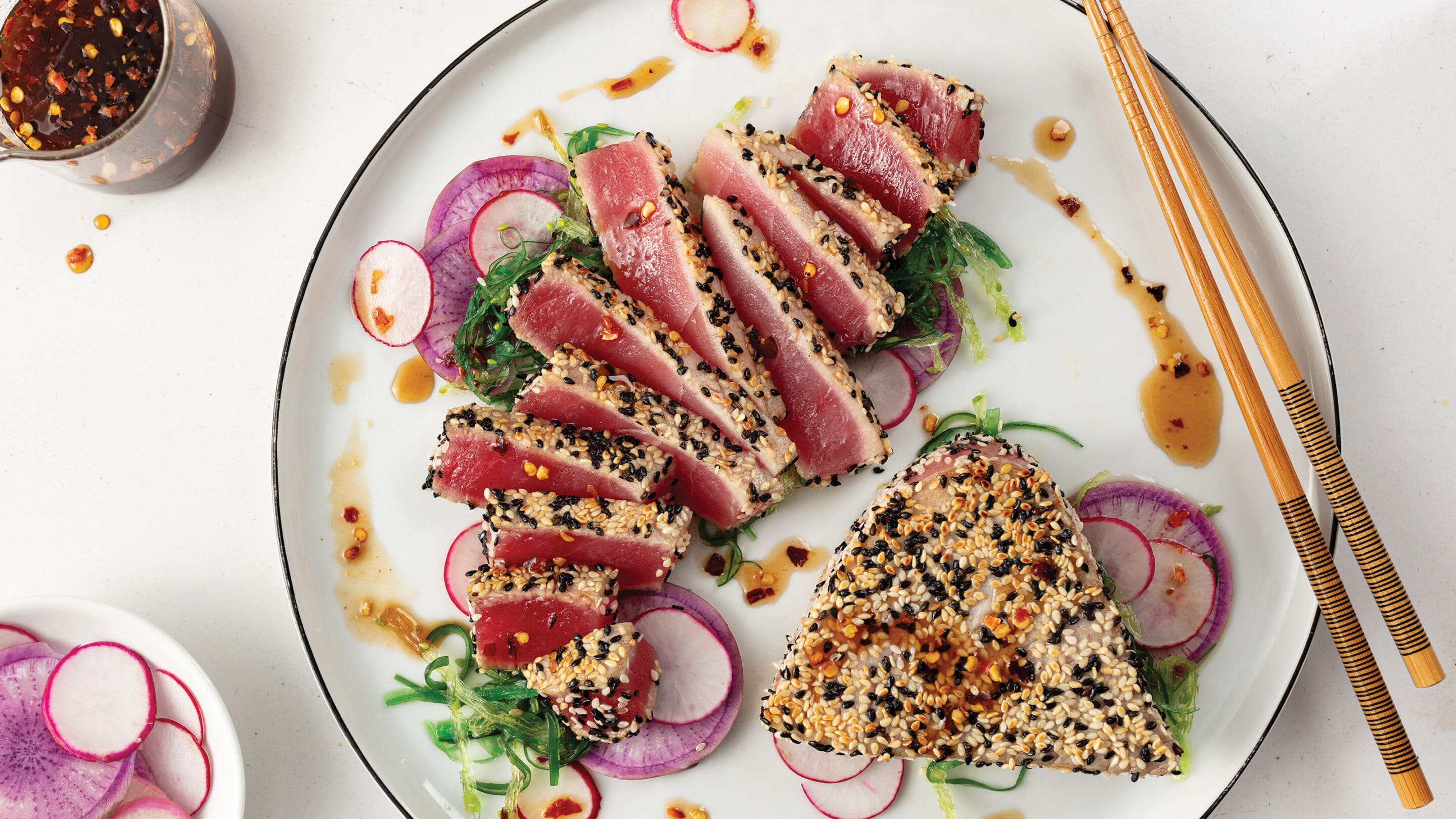 Two grilled sesame seed crusted ahi tuna steaks on white plate. One sliced medium-rare and expertly plated with red onion slices, seawood, and chili pepper sauce.