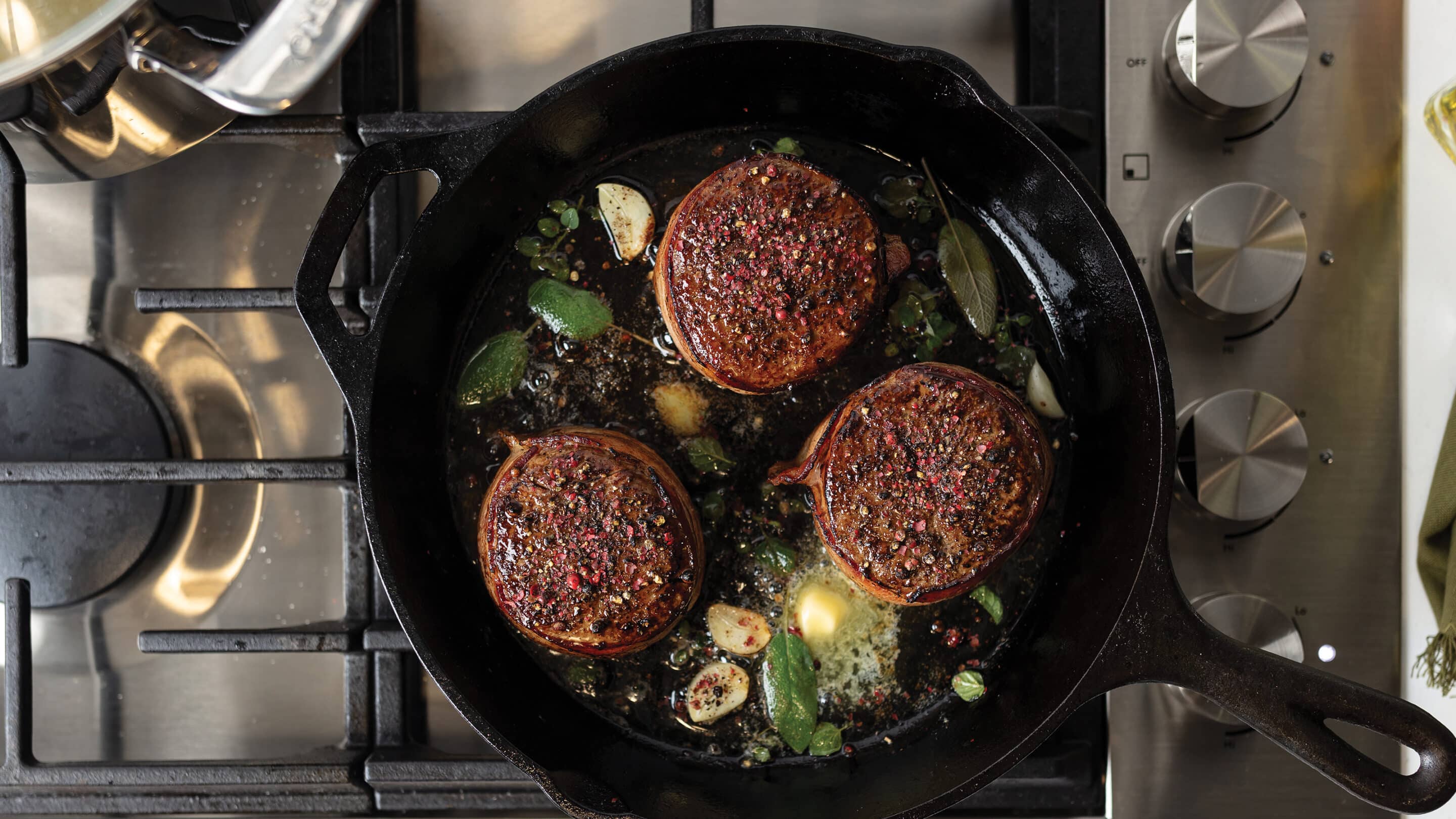 Three bacon-wrapped filet mignon steaks cooking in a cast iron skillet with garlic cloves, butter, and fresh herbs.