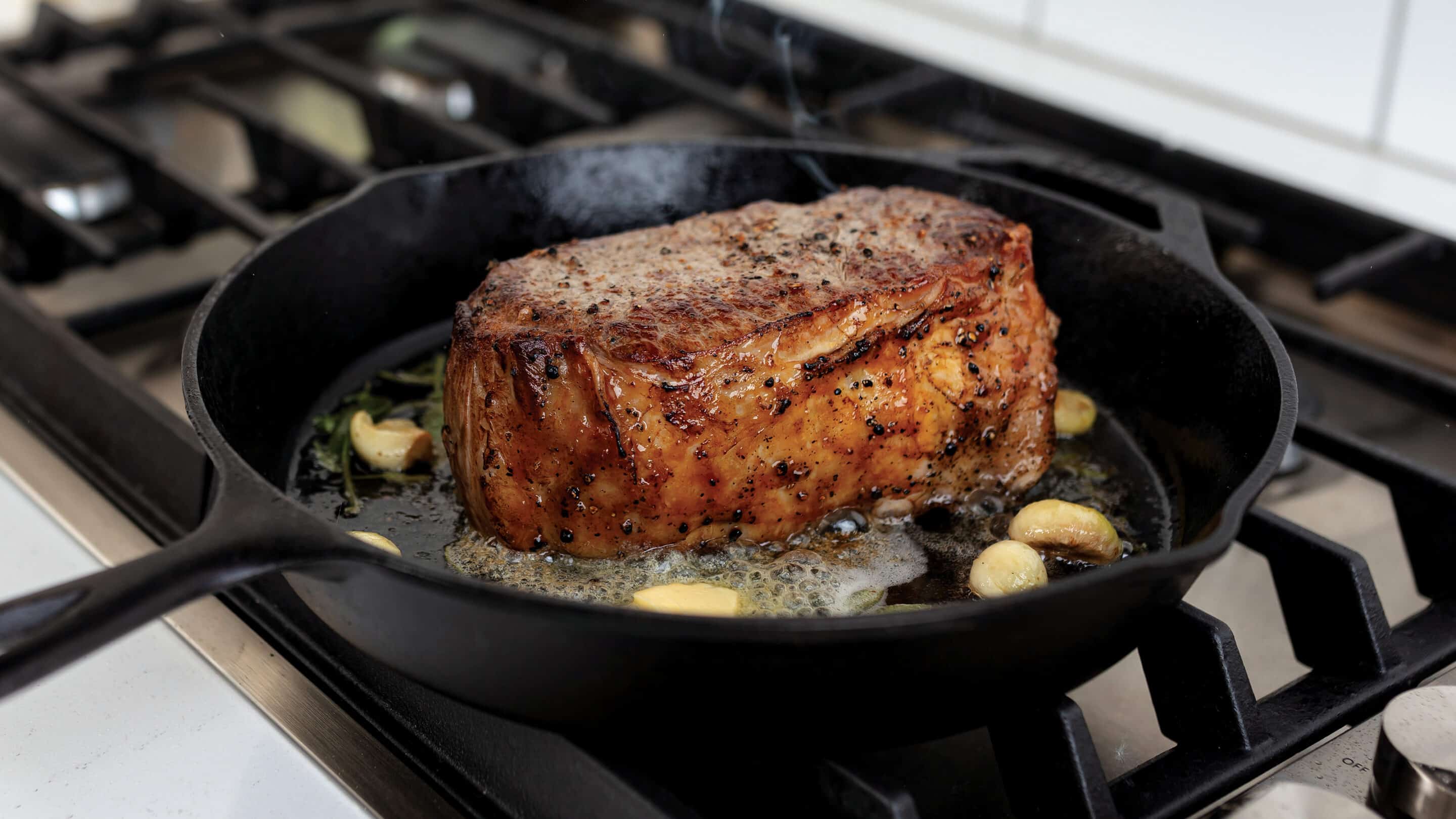 Seared King Cut New York strip steak in cast iron skillet with butter and garlic cloves on a gas stove.