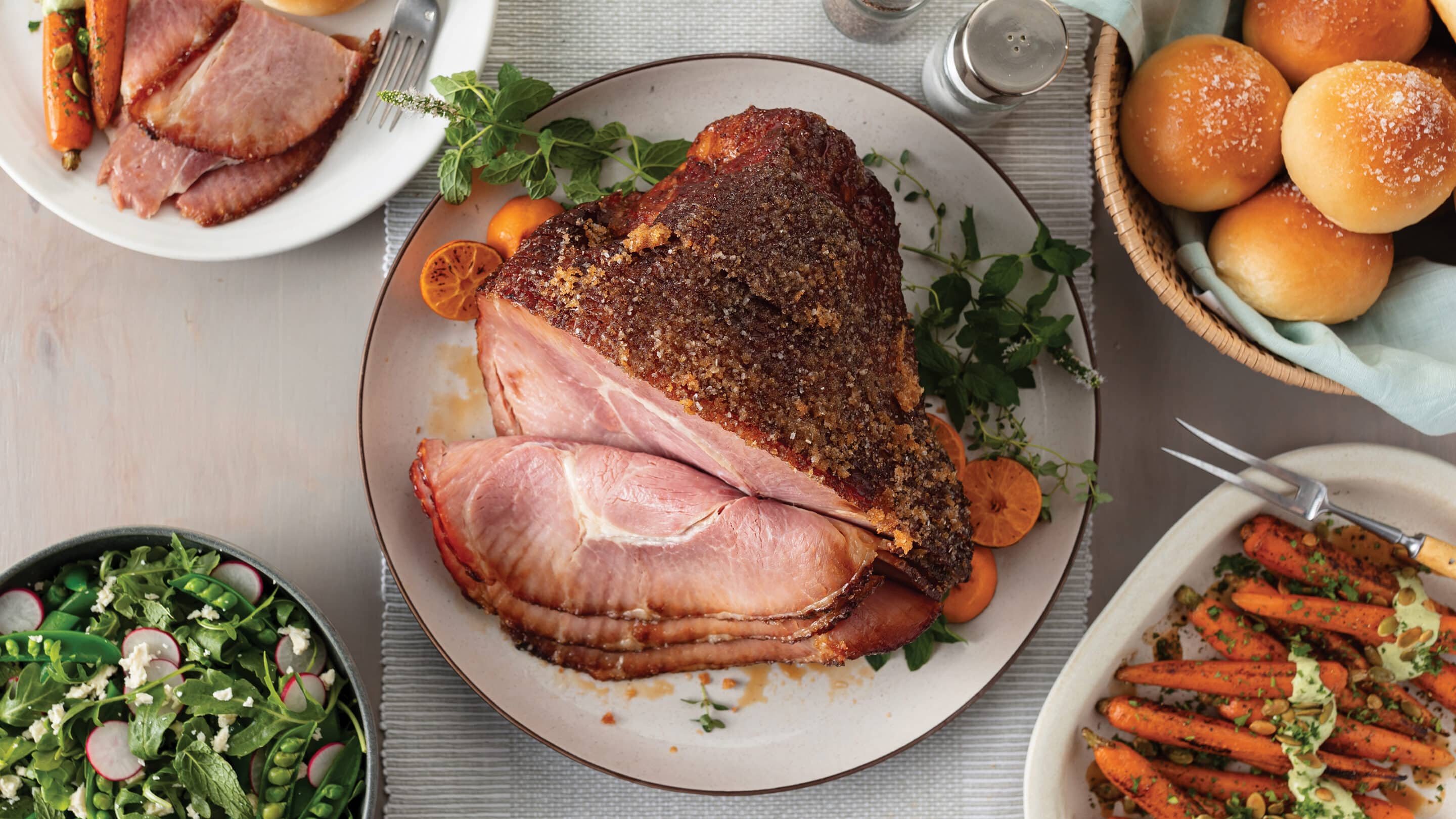 Table set with cooked spiral sliced ham, rolls, salad, and glazed carrots.