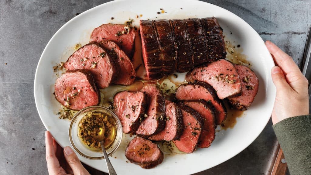 Person holding white platter with sear-roasted chateaubriand cooked medium-rare and sliced. Served with a small bowl of garlic and herb melted butter sauce.