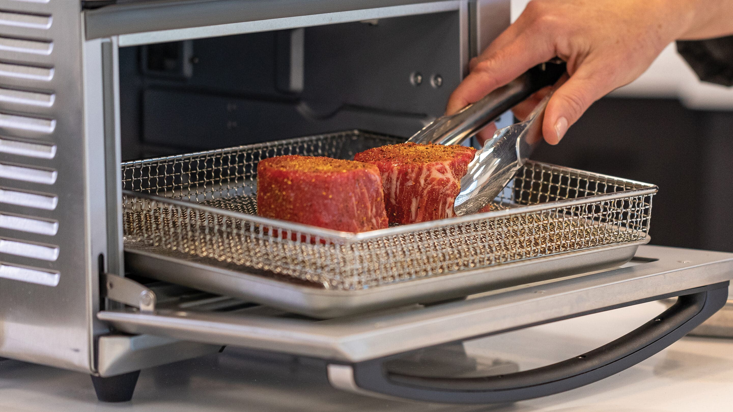 Chef using tongs to place a filet mignon steak in an air fryer next to another filet mignon.