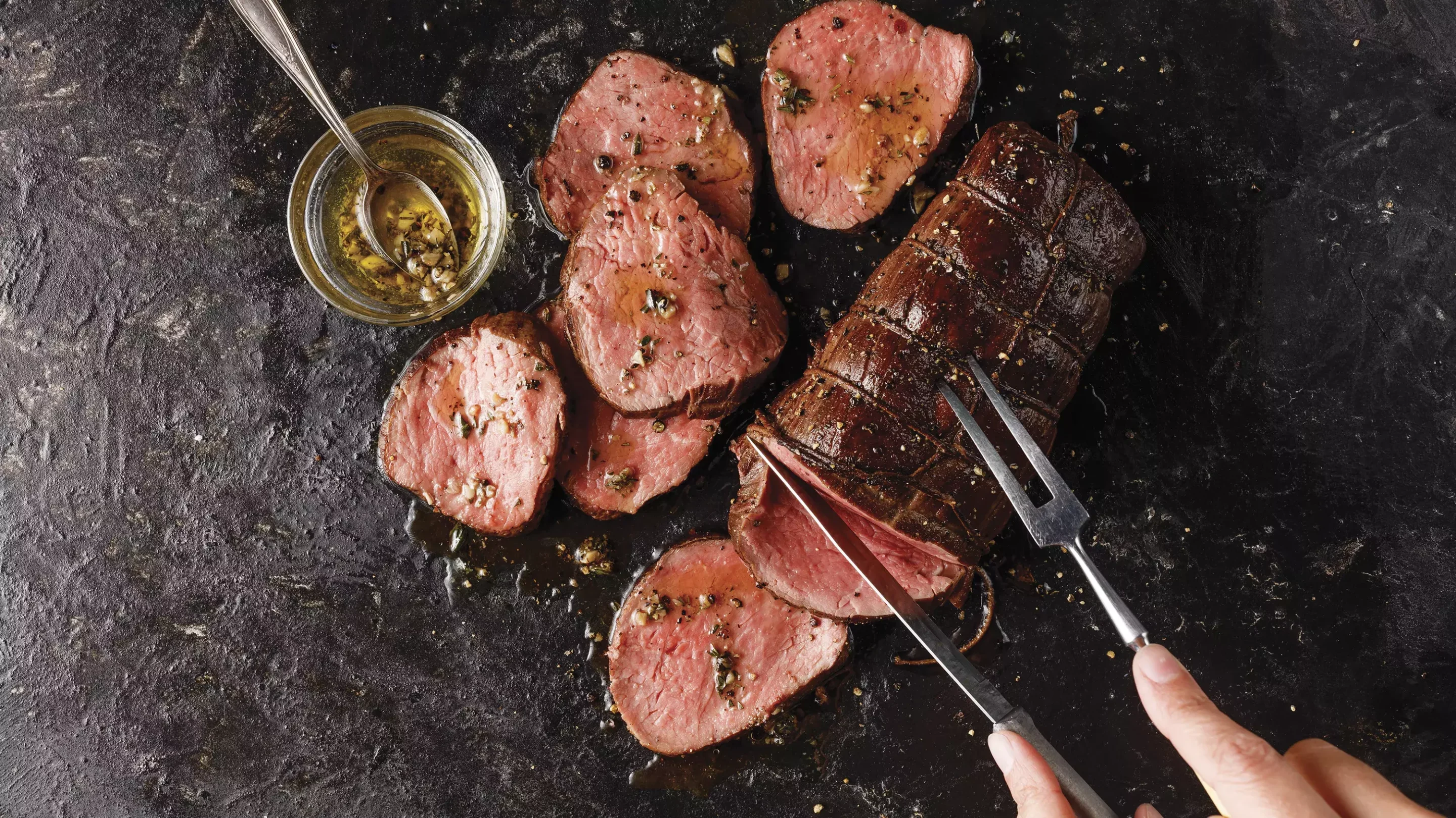 Three bacon-wrapped filet mignon steaks cooking in a cast iron skillet with garlic cloves, butter, and fresh herbs.