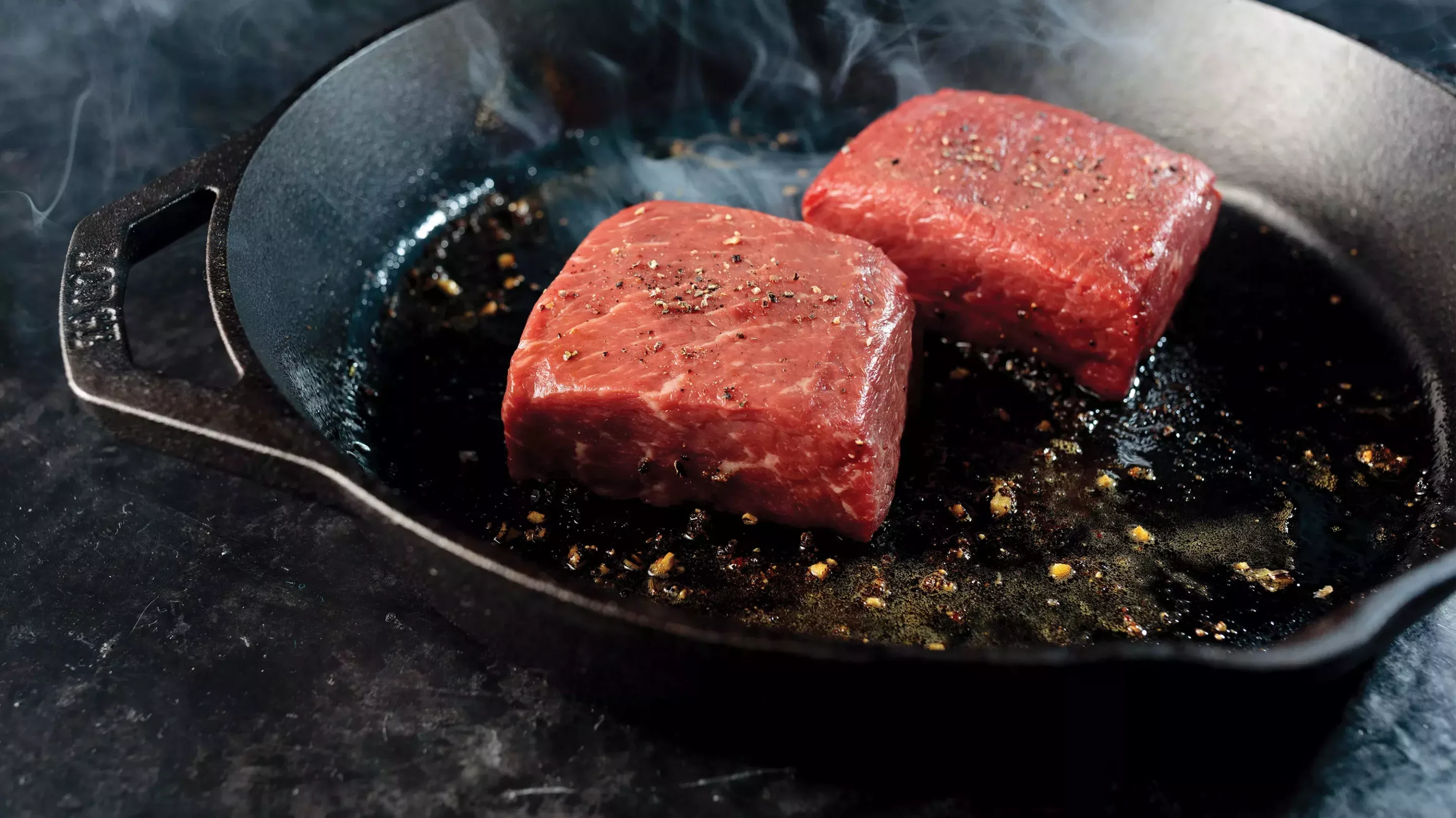 Three bacon-wrapped filet mignon steaks cooking in a cast iron skillet with garlic cloves, butter, and fresh herbs.