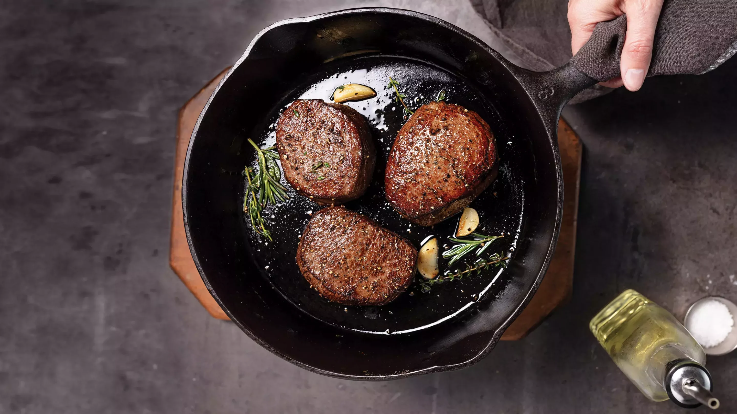 Three bacon-wrapped filet mignon steaks cooking in a cast iron skillet with garlic cloves, butter, and fresh herbs.