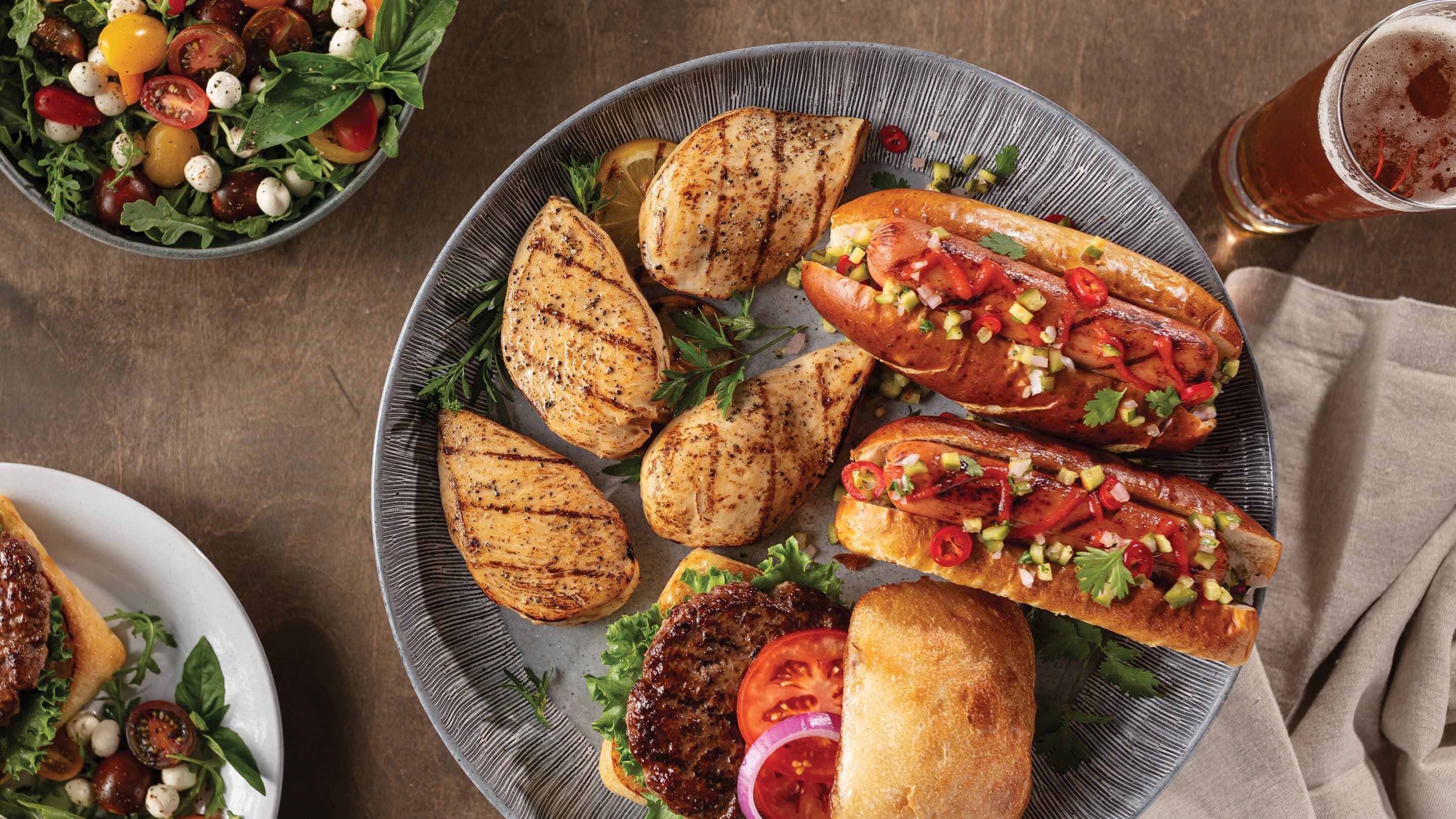 Large serving platter with 4 chicken breasts, 2 gourmet franks with toppings, burger with lettuce, tomato and red onion. Serving bowl with side salad. Smaller plate with burger and salad. Served with glass of water and glass of beer.