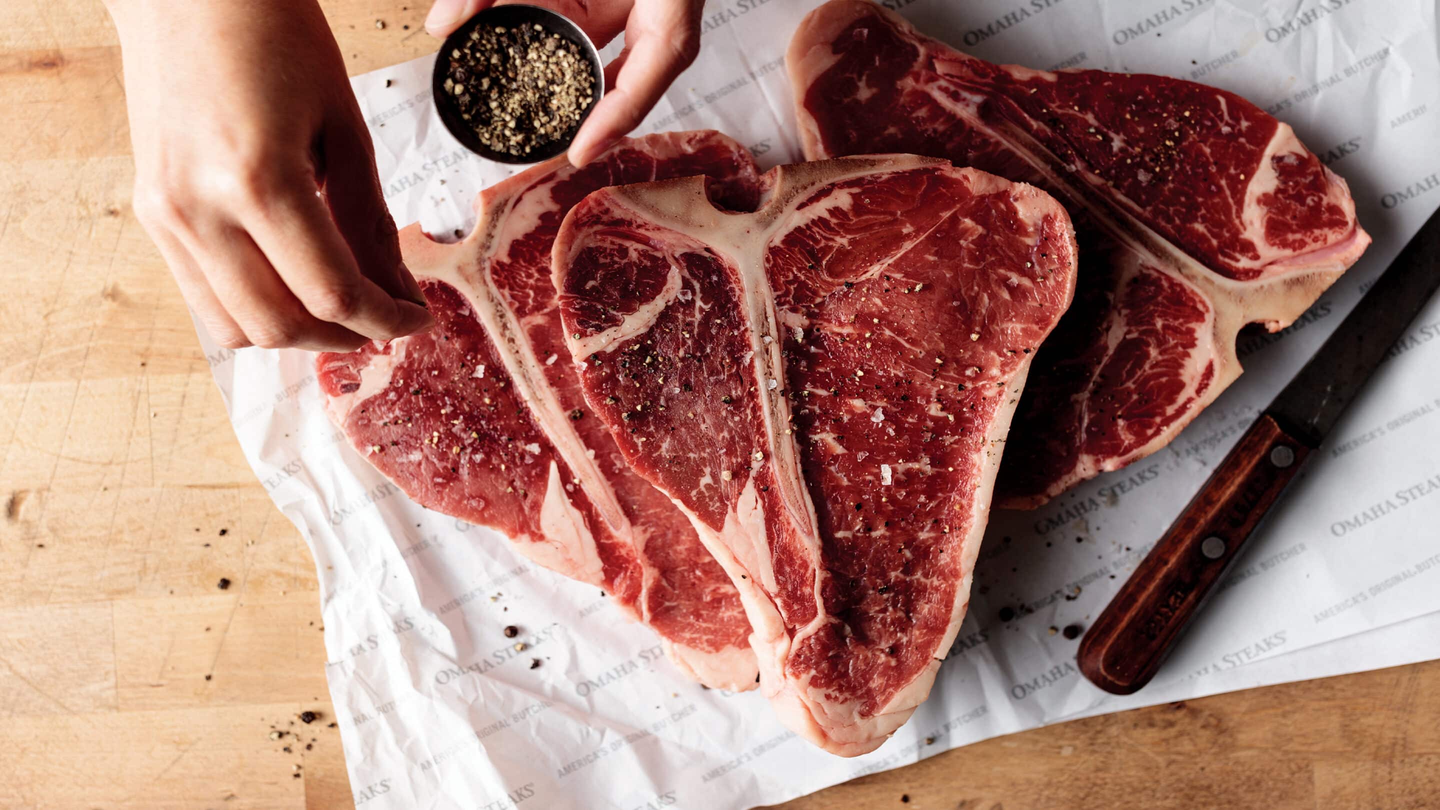 Person seasoning three raw t-bones setting on butcher's paper.