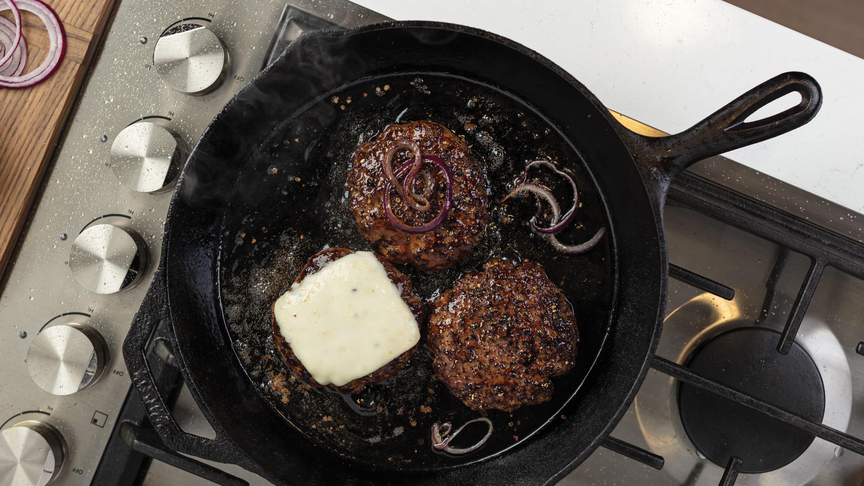 Cast iron skillet on stove with three pan-fried burgers, one with slice of white cheese cooking.