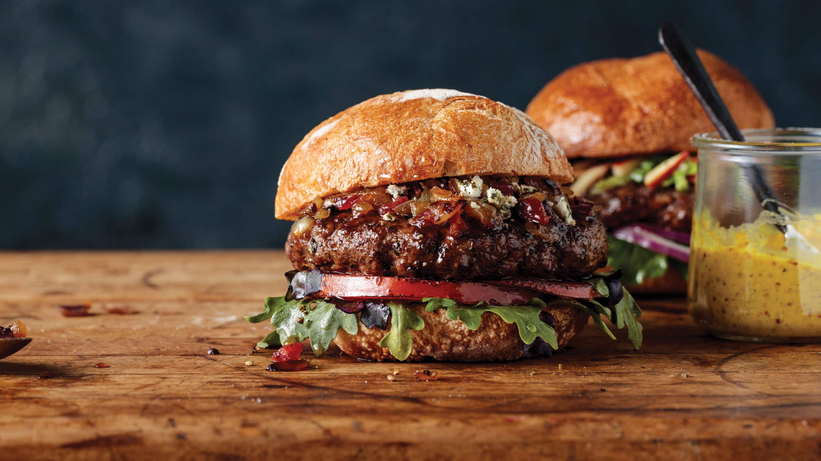 2 thick all-beef Omaha Steaks burgers with sauteed onions, lettuce, tomato, and sauce on a cutting board next to a jar of honey dijon mustard