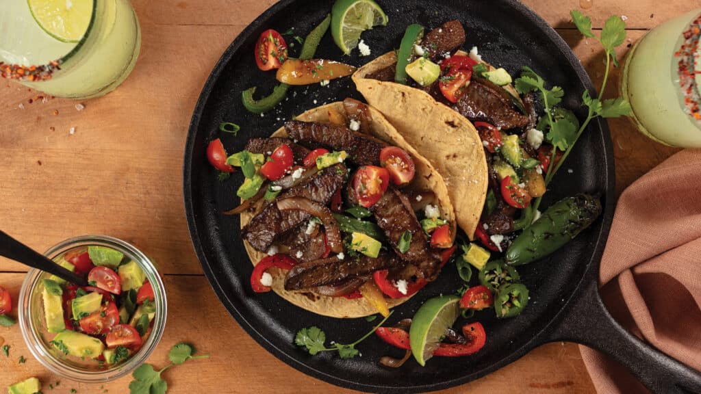 Cast iron skillet on wood counter with 2 fajitas filled with slices of flat iron steak, sliced jalapenos, a tomato and avocado salsa, cheese and cilantra.