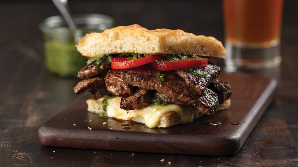 Sandwich on focaccia bread with sliced top sirloin steak, smoked mozzarella cheese, sliced plum tomato and pesto.