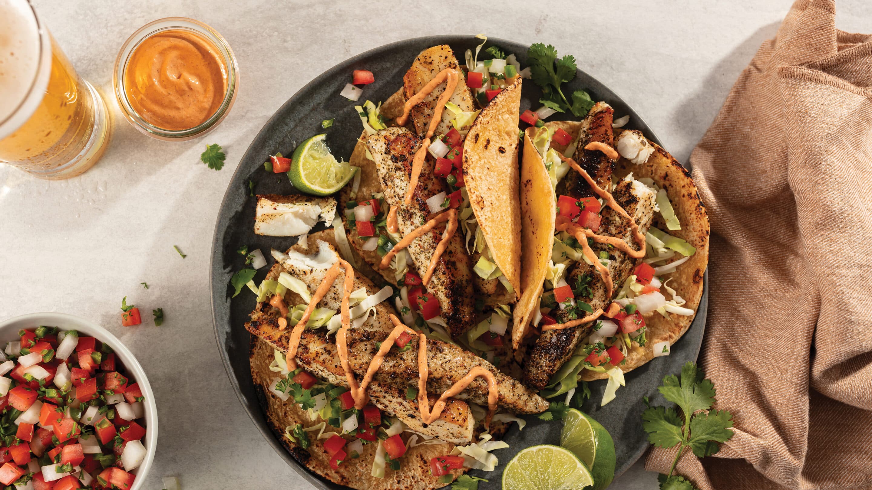 Three halibut filet tacos with fresh pico de gallo, lime wedges and cilantro and drizzled with chipolte mayonnaise plated next to a glass of light beer and bowl of pico de gallo.