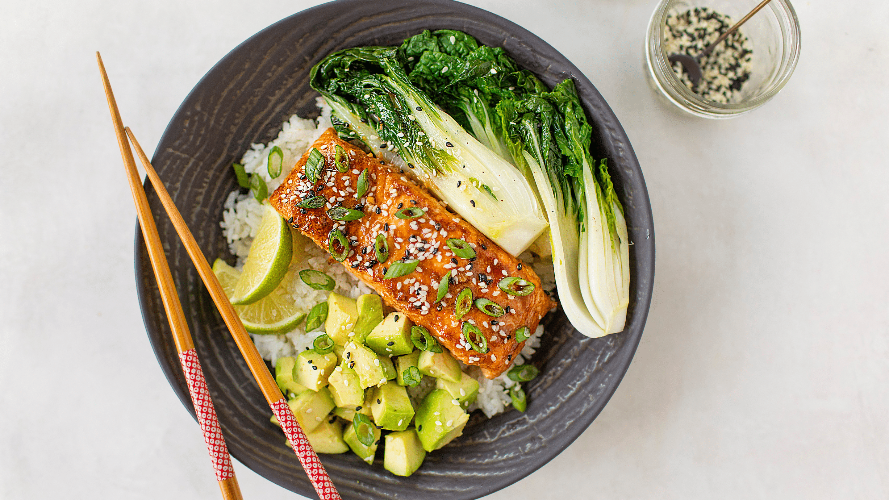 Teriyaki salmon bowl featuring Omaha Steaks salmon fillets, rice, bok choy, diced avocado, lime wedges in a grey bowl with chopsticks.