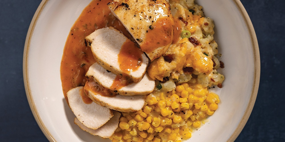 Pan-Seared Chicken With Cheesy Potatoes, Creamed Corn, and Creamy Tomato & White Wine Finishing Sauce on a white plate