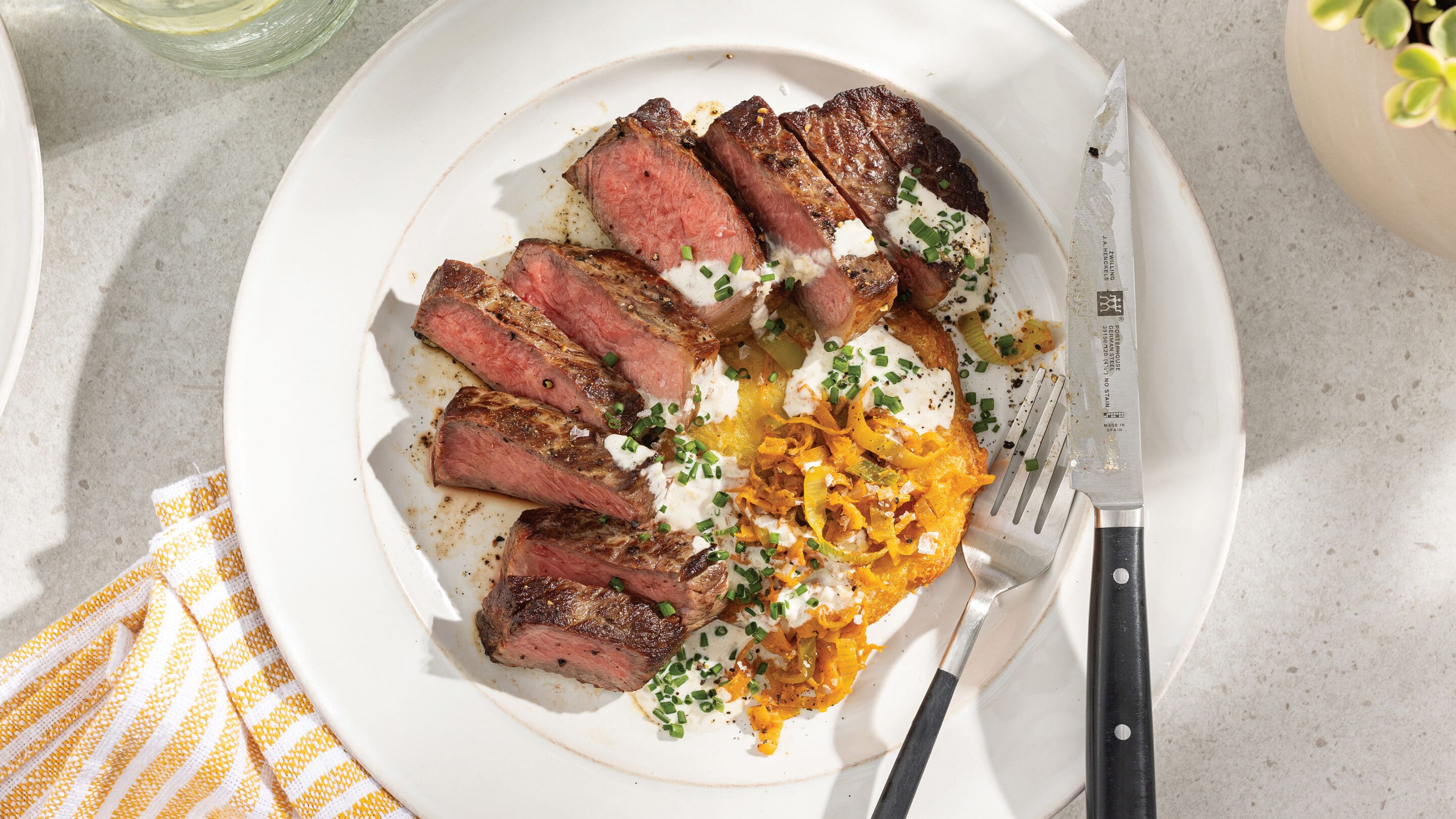 White plate on table with sliced New York strip steak cooked medium-rare and drizzled with garlic aioli and served with hash browns and melted leeks.